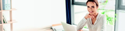 Dunkelhaarige Frau in weißer Bluse an einem Schreibtisch mit einem Laptop vor sich und lacht in die Kamera, während sie den rechten Arm aufstüzt und die Hand locker unter dem Kinn hält