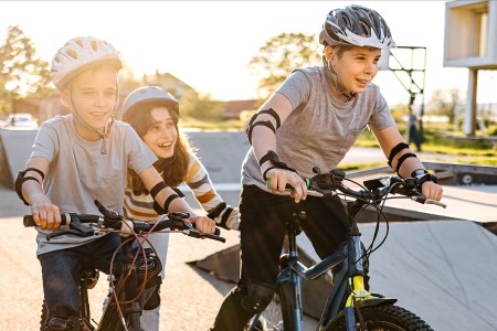 Zzwei Jungs und ein Mädchen, schätzungweise circa 10 bis 12 Jahre alt, fahren Fahrrad in der Nachmittagssonne und tragen alle Helme.