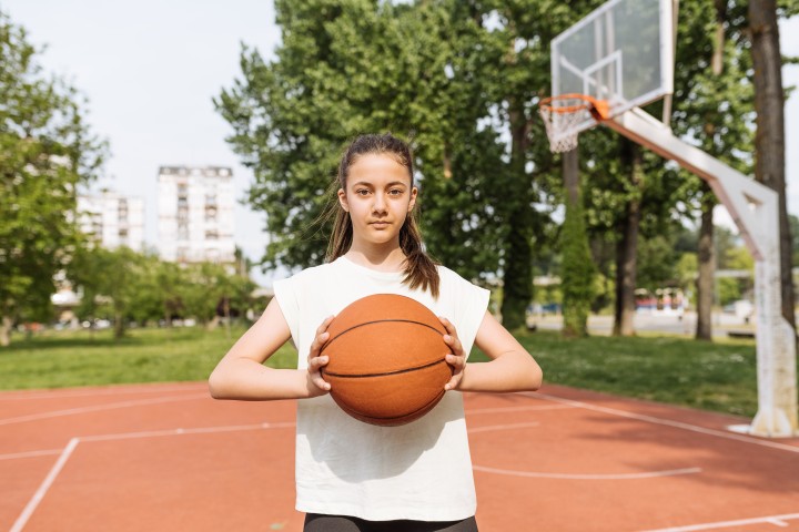 Teenagermädchen mit langen dunklen Haaren hält einen Basketbsll vor sich während sie in die Kamera schaut und unter dem Basketbsllkorb auf dem Basketballfeld steht