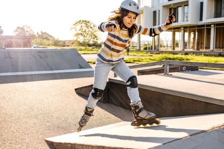 Ein Mädchen im Ringel-Pulli fährt mit Inlineskates in einer kleinen Halfpipe. Sie trägt einen grauen Helm und eine komplette Schutzausrüstung in schwarz. Die Sonne scheint im Hinterrgund ist ein modernes Mehrfamilienhaus.