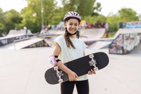 Teenagermädchen mit langen duknlen Haaren lacht in die Kamera und hat ein Skateboard unter dem Arm, im Hintergrund sind Halfpipes zu sehen