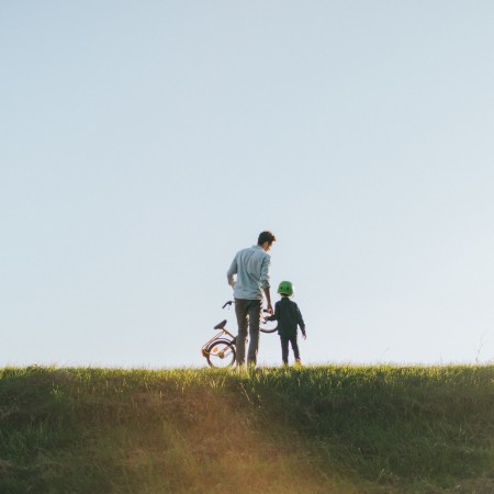 Weitläufige Wiese, auf dem weit hinten ein junger Vater mit seinem Sohn an der Hand von hinten zu sehen sind und der Vater trägt ein Kinderrad in der linken Hand, während die Abendsonne scheint