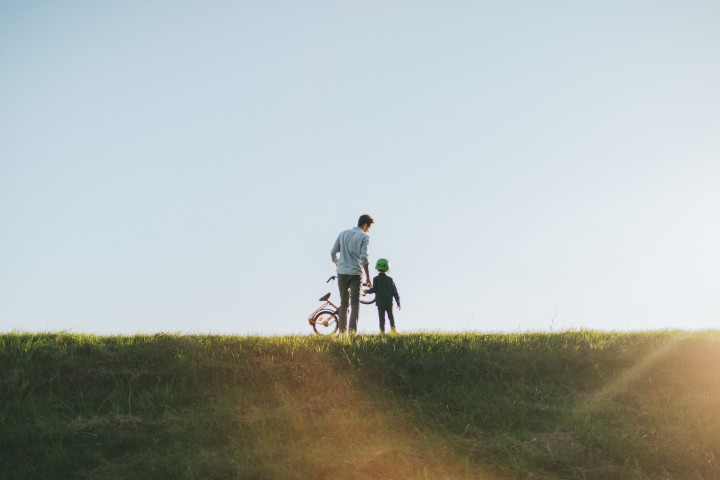 Weitläufige Wiese, auf dem weit hinten ein junger Vater mit seinem Sohn an der Hand von hinten zu sehen sind und der Vater trägt ein Kinderrad in der linken Hand, während die Abendsonne scheint