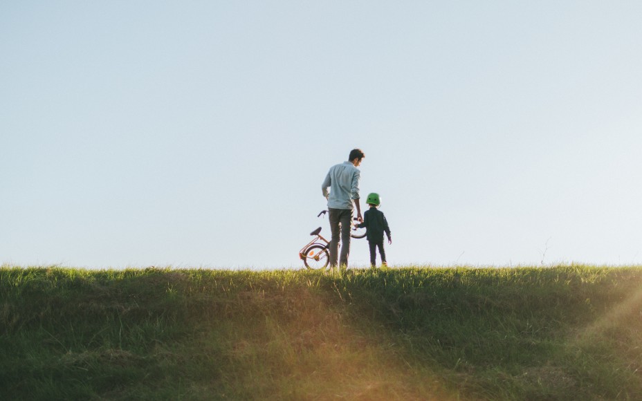 Weitläufige Wiese, auf dem weit hinten ein junger Vater mit seinem Sohn an der Hand von hinten zu sehen sind und der Vater trägt ein Kinderrad in der linken Hand, während die Abendsonne scheint