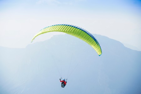 Mann in roter Jacke und Helm von hinten mit einem neongelben Gleitschirm, im Hintergrund nebelige Berge bei Sonnenschein und wolkenlos blauem Himmel