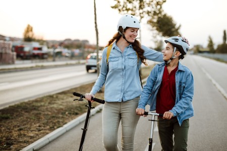 Mutter und Sohne schieben nebeneinander ihre E-Roller an einer Straße entlang, während sie sich anlachen, beide tragen einen Helm
