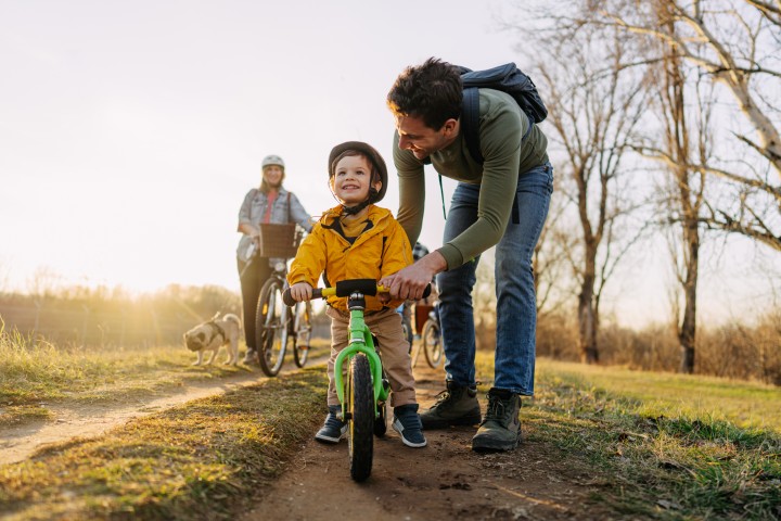 Vater stützt seinen Sohn, der auf einem grünen Kinderrad mit Helm auf einem Feldweg in der Abendsonne fährt