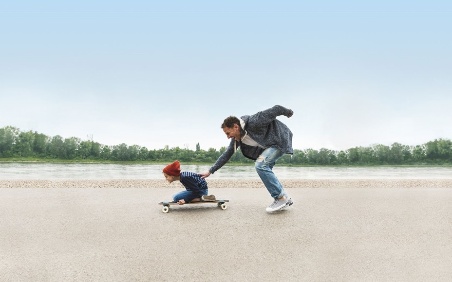 Vater schiebt auf Skateboard knienden Sohn an