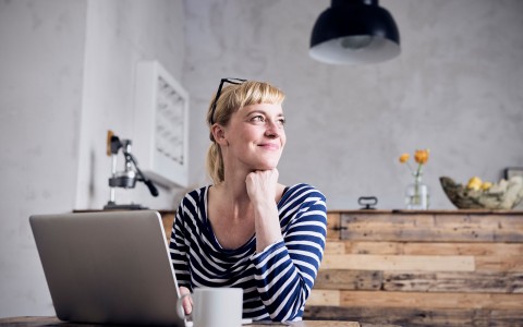Junge blonde Frau im blau-weißem Ringelshirt sitzt vor ihrem Laptop in einer Küche aus geflämmtem Holz mit einer Tasse und schaut lächelnd zur Seite