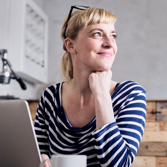 Junge blonde Frau im blau-weißem Ringelshirt sitzt vor ihrem Laptop in einer Küche aus geflämmtem Holz mit einer Tasse und schaut lächelnd zur Seite