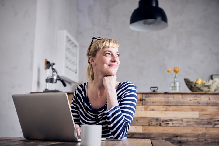 Junge blonde Frau im blau-weißem Ringelshirt sitzt vor ihrem Laptop in einer Küche aus geflämmtem Holz mit einer Tasse und schaut lächelnd zur Seite