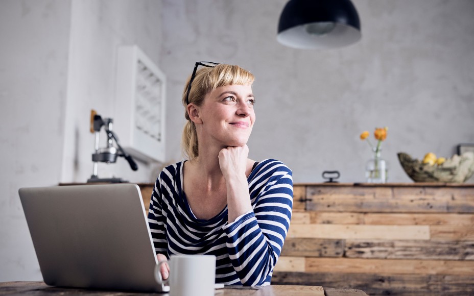 Junge blonde Frau im blau-weißem Ringelshirt sitzt vor ihrem Laptop in einer Küche aus geflämmtem Holz mit einer Tasse und schaut lächelnd zur Seite