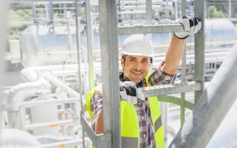 Mann mit weißem Bauhelm und gelber Sicherheitsweste auf einem Baugerüst lächelt entspannt in die Kamera