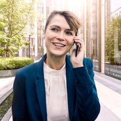 Lachende Frau mit verschränkten Armen telefoniert, während sie draußen vor einem Bürogebäude und einer großen Fensterfront steht.