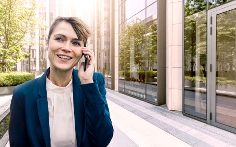 Lachende Frau mit verschränkten Armen telefoniert, während sie draußen vor einem Bürogebäude und einer großen Fensterfront steht.