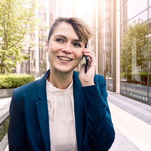 Lachende Frau mit verschränkten Armen telefoniert, während sie draußen vor einem Bürogebäude und einer großen Fensterfront steht.