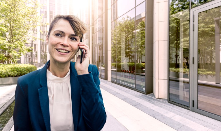 Lachende Frau mit verschränkten Armen telefoniert, während sie draußen vor einem Bürogebäude und einer großen Fensterfront steht.