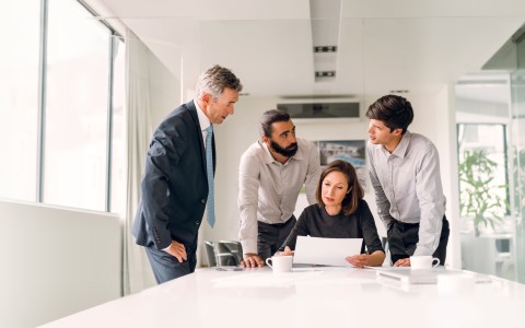 Drei Businessmänner umstehen an einem Konferenztisch eine sitzende Kollegin und alle schauen auf eine Papier, das die Frau in den Händen hält