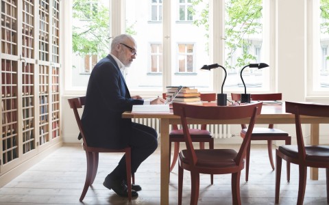 Steeurberater oder Wirtschaftsprüfer sitzt in einem Altbau-Büro an seinem Schreibtisch, auf dem viele Bücher liegen, und schreibt etwas, links sieht man eine komplett Wand mit Vitrinenschränken voller Akten