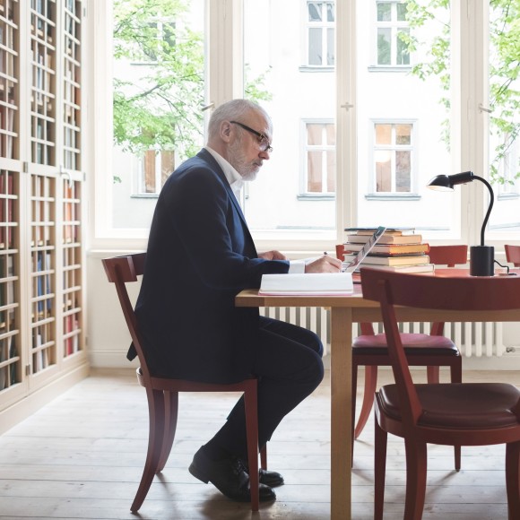 Steeurberater oder Wirtschaftsprüfer sitzt in einem Altbau-Büro an seinem Schreibtisch, auf dem viele Bücher liegen, und schreibt etwas, links sieht man eine komplett Wand mit Vitrinenschränken voller Akten