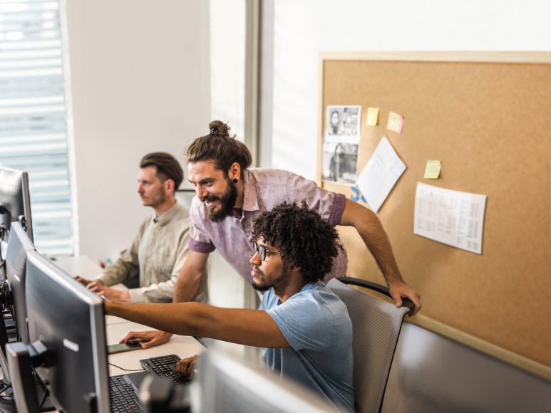 Mänenr in einem Büro, von denen zwei vor Bildschirmen sitzen und einer beugt sich ovn hinten über die Schulter eines der Männer und zeigt lachend auf den einen Bildschirm