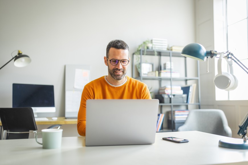 Mann mit Dreitagbart und Brille im orangefarbenen Pullover sitzt in einem Büro an seinem Schreibtisch vor dem Laptop