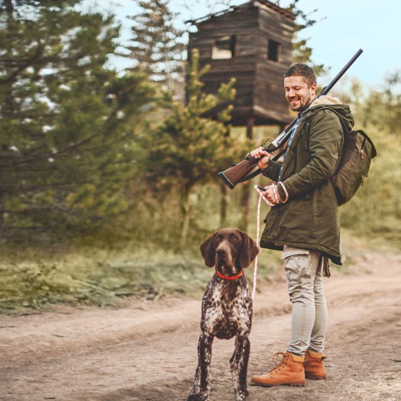 Lächelnder Jäger steht in typischer Jagdkleidung vor einem Hochsitz im Wald, hat sein Gewehr geschultert und seinen großen Jsgdhund an der Leine