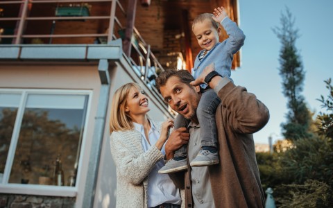 Glücklich lachende Familie vor ihrem Haus und der Vater trägt den kleinen blonden Sohn auf den Schultern
