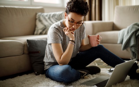 Junge Frau mit kurzen dunklen Locken und Brille sitzt im Schneidersitz auf dem Sofa vor ihrem Laptop und schaut nachdenklich darauf