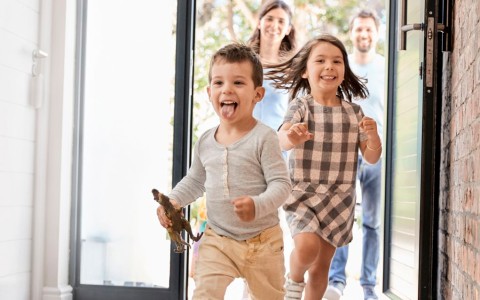 Kinder, ein Mädchen und ein Junge stürmen lachend durch die Haustür in den Hausflur, der Junge streckt die Zunge heraus und hält einen Dino in der Hand, hinter den beiden folgen die Eltern ebenfalls lachend