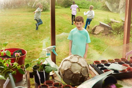 Junge schaut entsetzt auf eine zerbrochene Fensterscheibe, hinter der ein alter Lederfußball liegt, während zwei andere Kinder über eine Rasen davonlaufen
