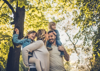 Junge Familie glücklich im Wald bei Sonnenschein, Mutter trägt die Tochter Huckepack, der Vater hat den Sohn auf den Schultern