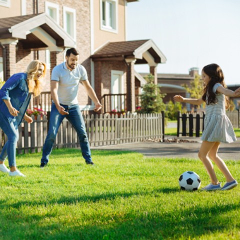 Eltern spielen mit ihrer Tochter auf der Wiese vor zwei geklinkerten Reihenhäusern Fußball in der Sonne