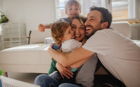 Junge Familie mit zwei kleinen Kindern umarmen sich alle glücklich lachend und knuddeln im Wohnzimmer