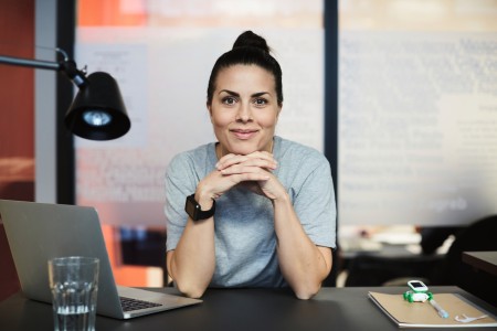 Junge Frau mit Dutt schaut in die Kamera und stützt das Kinn auf ihre auf dem Schreibtisch aufgestützen Hände, neben ihr steht ein Laptop