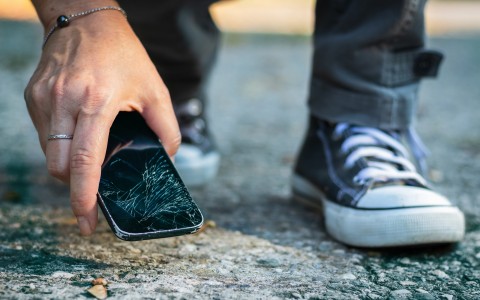 Junger Mann, von dem man nur die Füße mit dunklen Converse sieht, hebt ein heruntergefallenes Handy mit gesplittertem Display vom Asphalt auf