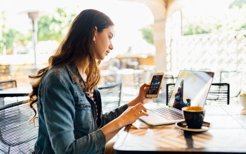 Frau mit langen dunklen Haaren schaut konzentriert auf ihr Handy in der linken Hand mit vermutlich Aktienkursen während sie mit der anderen Hand auf dem Laptop tippt