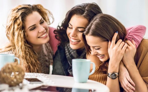 Drei Freundinnen sitzen lachend an einem Cafehaus-Tisch, liegen sich in den Armen und haben Tassen vor sich stehen