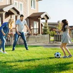 Eltern spielen mit ihrer Tochter auf der Wiese vor zwei geklinkerten Reihenhäusern Fußball in der Sonne