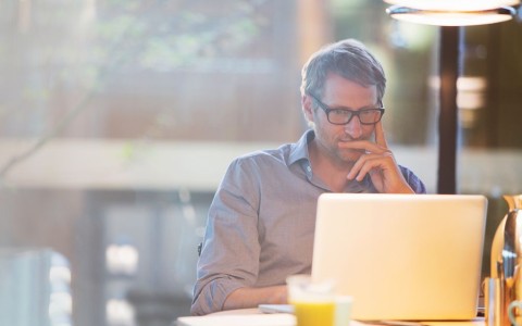Mann mit Brille sitzt nachdenklich vor seinem Laptop und hat die Hand am Kinn abgestützt