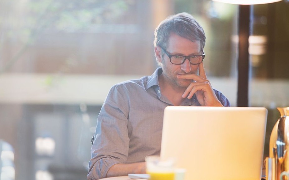 Mann mit Brille sitzt nachdenklich vor seinem Laptop und hat die Hand am Kinn abgestützt