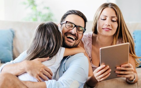 Vater mit Bart sitzt vor einem hellen Sofa auf dem Boden und lacht während sich seine Tochter ihre Arme um seinen Hals schlingt und er sie festhält. Seine Frau liegt bäuchkings hinter ihm mit einem Tablet, auf das sie beide schauen
