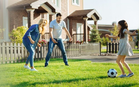 Eltern spielen mit ihrer Tochter auf der Wiese vor zwei geklinkerten Reihenhäusern Fußball in der Sonne