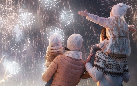Von hinten zu sehende Familie mit zwei kleinen Kindern schaut einem großen Feuerwerk in Weiß- und Goldtönen zu, der Vater hat die ältere Tochter auf den Schultern, die Mutter trägt das kleinere Kind auf der Hüfte