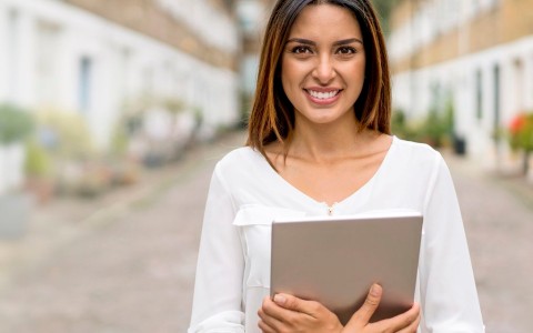 Frau mit langen Haaren hält ihren Laptop im Arm und steht auf einer Straße, die auf beiden Seiten von einer langen Reihe weißer Reihenhäuser gesäumt ist