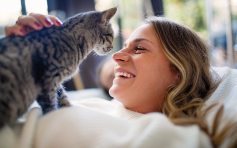 Blonde Fru liegt uf dem Sofa und strahlt ihre grau getigerte Katze an, die auf ihrer Brust steht