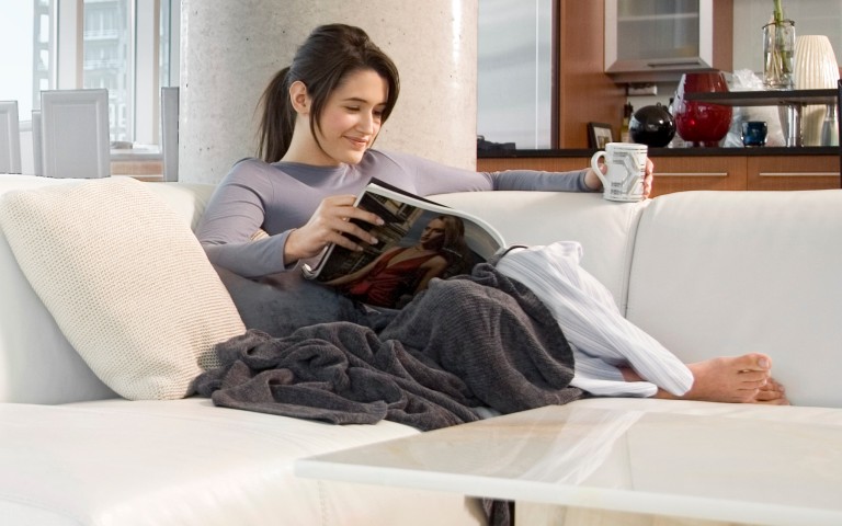Dunkelhaarige Frau sitzt auf einem hellen modernen Sofa in eine Decke gekuschelt mit einer tasse in der linken Hand und liest in einer Zeitschrift