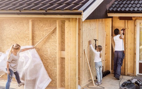 Familie mit blonder Tochter verkleidet ein haus von außen mit neuen Spanplatten, Tochter hämmert etwas und der Vater schraubt mit einem Akkuschrauber.