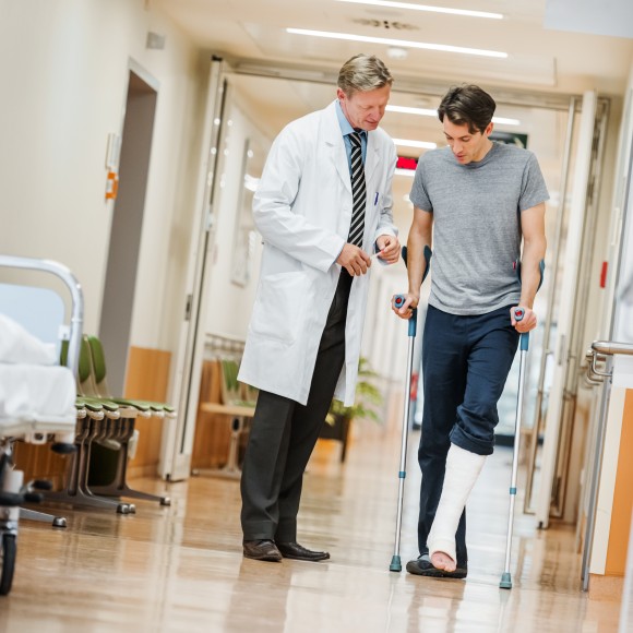 Ein Mann mit eingegipstem Fuß und Krücken steht in einem Krankenhauskorridor und redet mit einem Arzt
