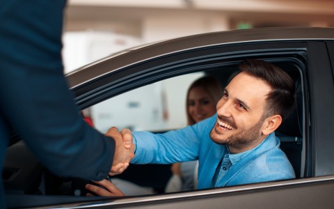 Mann sitzt lachend in einem Auto und schüttelt einem anderen Mann durch das Fahrerfenster die Hand